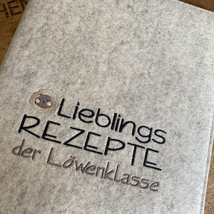 May include: A light gray felt recipe book cover with embroidered text and a lion illustration. The text reads "Lieblings REZEPTE der Löwenklasse" in black and gray thread. The lion is tan and brown.
