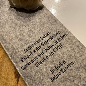 Può includere: Una pochette in feltro grigio con il testo "Liebe das Leben, Erlaube dir Schwächen, Vertraue auf deine Stärken, Glaube an DICH, In Liebe, deine Eltern" ricamato in nero.