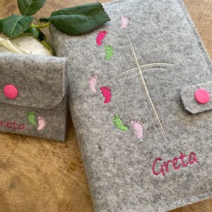 May include: Gray felt Bible cover with a white cross and pink and green footprints embroidered on it. The cover has a flap with a pink button and the name "Greta" is embroidered on the bottom.