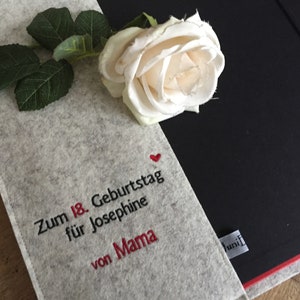 May include: A gray felt notebook cover with embroidered text and a red heart, paired with a white rose and green leaves. The text reads "Zum 18. Geburtstag f&uuml;r Josephine von Mama."