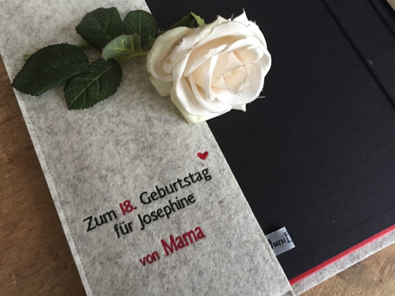 May include: A gray felt notebook cover with embroidered text and a red heart, paired with a white rose and green leaves. The text reads "Zum 18. Geburtstag f&uuml;r Josephine von Mama."