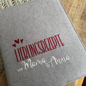Könnte beinhalten: Graues Filz-Rezeptbuch mit dem gestickten Text "Lieblingsrezepte von Mama für Anna" in Rot und Weiß.