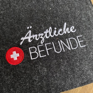 Könnte beinhalten: Ein grauer Filzordner mit weiß gesticktem Text, der "ärztliche BEFUNDE" lautet, und einem roten Kreis mit einem weißen Kreuz in der linken unteren Ecke.