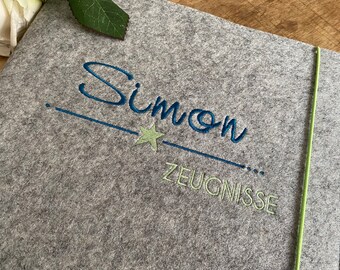 Könnte beinhalten: Grauer Filzordner mit grünem Gummibandverschluss. Der Ordner ist mit dem Namen "Simon" in blauer Farbe, einem grünen Stern und dem Wort "Zeugnisse" in grüner Farbe bestickt.