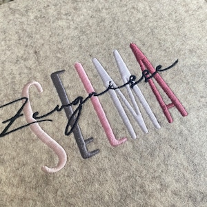 Könnte beinhalten: Eine personalisierte Filz-Aufbewahrungsbox mit dem Namen "Selma", der in mehreren Farben gestickt ist, darunter Rosa, Grau und Rot.