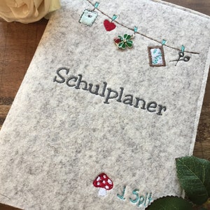 May include: A light gray felt planner cover with embroidered details. The cover features the text "Schulplaner" and a whimsical design of hanging objects. A red mushroom and the name "J. Spilt" are also embroidered on the cover.