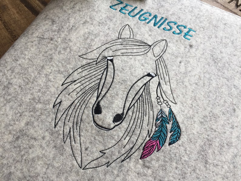 May include: Embroidered design of a horse's head with feathers and beads on a gray felt background. The text "ZEUGNISSE" is embroidered in blue above the horse.