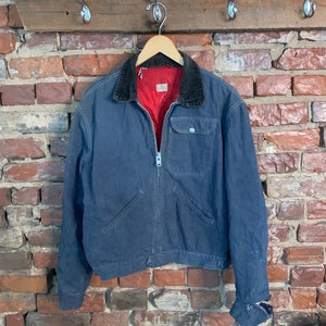 Op de afbeelding: Een vintage blauwe werkjas met een corduroy kraag en een rode gewatteerde voering. De jas heeft een ritssluiting, twee voorzakken en een manchet met knoop. Hij hangt aan een houten hanger tegen een bakstenen muur.