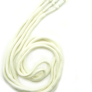 Puede incluir: Un conjunto de cuatro cordones trenzados tubulares de color blanco roto dispuestos en una bobina suelta. Cada cordón presenta una punta cilíndrica de plástico blanco sólido en un extremo, diseñada para pasar a través de ojales o canales.