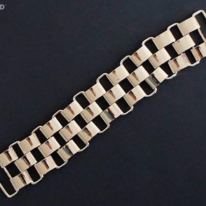 Puede incluir: Una cadena de metal dorado con un patrón de eslabones rectangulares. La cadena tiene dos bucles en cada extremo.