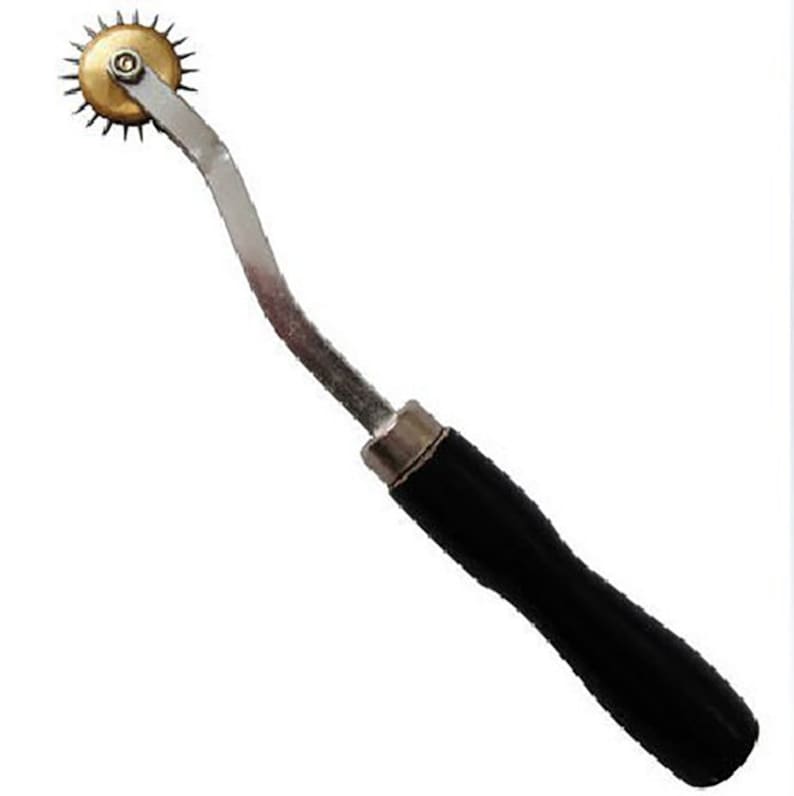 May include: A silver and brass sewing tool with a black handle. The tool has a circular wheel with sharp points and a curved handle.