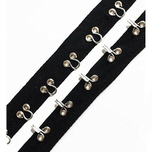May include: Two strips of black fabric with evenly spaced silver hook and eye closures. The hooks are on one strip, and the eyes are on the other. The background is white.