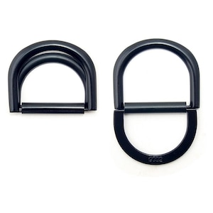 Könnte beinhalten: Zwei schwarze Metall-D-Ringe. Einer ist ein Doppelring, der andere ein Einzelring mit horizontaler Stange. Die Ringe haben eine matte Oberfläche und sind vor weißem Hintergrund platziert. Auf dem Einzelring ist die Nummer "9002" eingraviert.