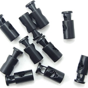 Puede incluir: Diez cierres de cordón de plástico negro con un mecanismo de resorte. Los cierres de cordón son de forma cilíndrica con un pequeño agujero en la parte superior y un agujero más grande en la parte inferior.