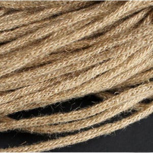 May include: Close-up of a coil of thick, natural-colored jute rope. The rope is tightly woven with a textured surface, showing individual fibers. The neutral color and robust texture suggest its use in crafts, decor, or other projects.