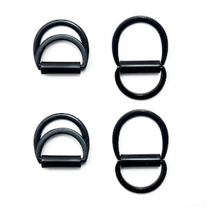 Pode incluir: Quatro argolas em D de metal preto com acabamento fosco dispostas sobre um fundo branco. Duas argolas são em forma de duplo D e duas em D simples. As argolas são conectadas por uma barra cilíndrica.