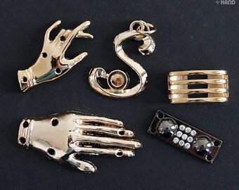 May include: Five gold-toned metal charms, including two hands, a letter S, a rectangular shape with three horizontal lines, and a black and silver rectangular shape with small white dots.