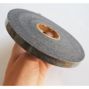 May include: A roll of gray double-sided tape. The tape is held in a hand and has a brown cardboard core. The tape is used for crafts, scrapbooking, and other projects.