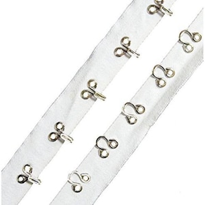 May include: Close-up of white fabric strips with silver metal hook and eye closures. The closures are evenly spaced along the fabric. The hooks are looped, and the eyes are figure-eight shaped.