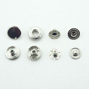 May include: A collection of eight silver-colored snap fasteners. The fasteners are in various stages of assembly, with both the top and bottom pieces visible. The metal has a shiny, reflective surface.