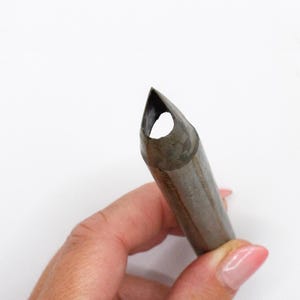 Könnte beinhalten: Nahaufnahme eines Metall-Lederstanzwerkzeugs. Das Werkzeug ist zylindrisch mit einem spitz zulaufenden Ende und einem Loch. Das Werkzeug wird von einer Hand vor weißem Hintergrund gehalten. Das Werkzeug ist dunkelgrau.