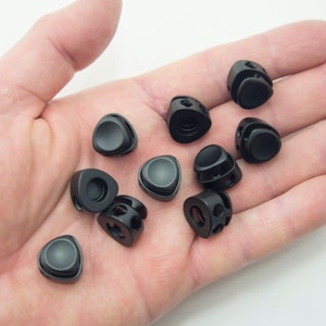 以下が含まれることがあります： 三角形の黒いプラスチック製のトグルを手に持った手のアップ。トグルは中央に穴があり、両側に小さな穴があります。