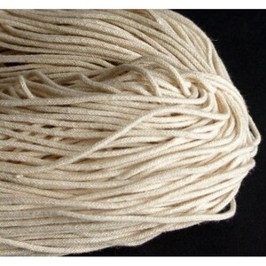 May include: A close-up of a bundle of off-white cotton rope. The rope is tightly wound, with visible strands and a slightly textured surface. The neutral color suggests a natural fiber, suitable for various crafting or decorative purposes.
