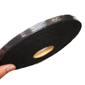 May include: A roll of black tape with a light-colored, patterned edge. The tape is tightly wound, revealing a central cardboard core. The image is shot at an angle, highlighting the tape's texture and form.