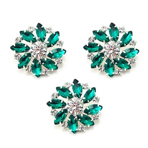 Puede incluir: Tres adornos florales decorativos con un brillante central transparente rodeado de cristales verdes esmeralda en forma de marquesa y brillantes transparentes más pequeños. Los adornos tienen un reverso de metal plateado sobre fondo blanco.