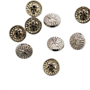 以下が含まれることがあります： 銀色の金属製スナップファスナーのコレクション。シンプルな円形デザインのものもあれば、テクスチャ加工された装飾的な模様のものもあります。ファスナーは白い背景に散らばっています。