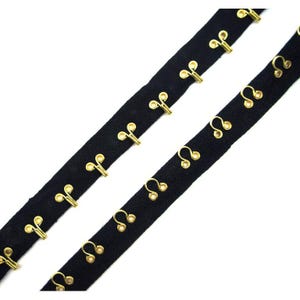May include: Two strips of black hook and eye tape with gold-coloured metal closures. The hooks and eyes are evenly spaced along the black fabric. The tape is angled diagonally across a white background, showcasing the sewing notions.