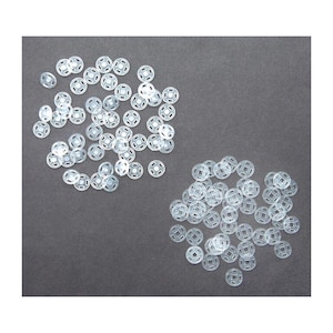 Könnte beinhalten: Eine Ansammlung klarer Kunststoff-Druckknöpfe, verstreut auf einer dunkelgrauen Oberfläche. Die Druckknöpfe sind rund mit einem zentralen Loch und einer Reihe kleinerer Löcher am Umfang. Diese Verschlüsse werden häufig in Näh- und Bastelprojekten verwendet.