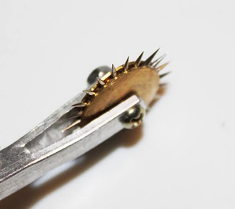 May include: A close-up of a silver tool with a brass wheel with sharp points. The wheel is attached to the tool with a screw.
