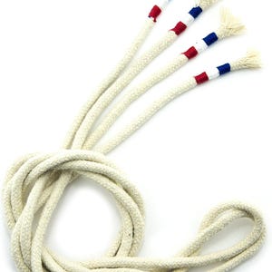 May include: A set of four off-white braided cotton cords coiled at the base. Each cord features a decorative tip wrapped in red, white, and blue thread, ending in a frayed, unraveled tassel. The cords are uniform in texture and thickness.