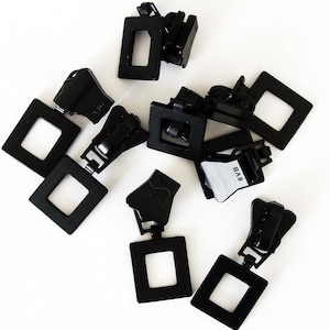 May include: A collection of black metal zipper pulls featuring a square-shaped design. Each pull has a hinged mechanism and a rectangular opening in the center. Some of the components are marked with the text 8VB on the side.