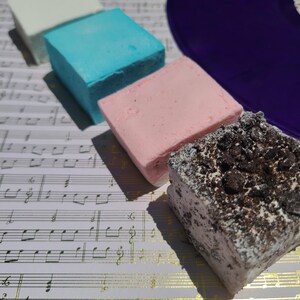 May include: Four square-shaped marshmallows arranged in a diagonal line. The first is white, the second is bright blue, the third is light pink, and the fourth is white with a dark brown cookie crumble topping. A purple circular object is visible.