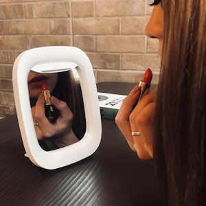 May include: A white lighted vanity mirror with a round shape and a woman applying red lipstick in the reflection.