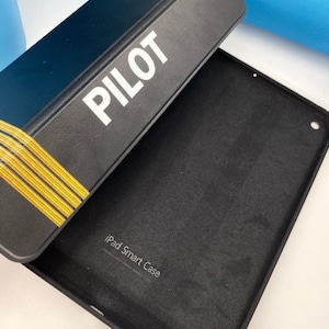 May include: Black iPad Smart Case with the word "PILOT" printed in white on the front. The case is open and the inside is lined with a soft black material.