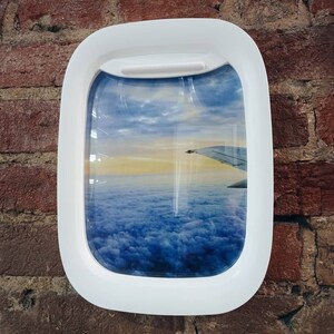 K&ouml;nnte beinhalten: Ein wei&szlig;er Flugzeugfensterrahmen mit Blick auf Wolken und einen Flugzeugfl&uuml;gel in der Ferne. Die Wolken sind wei&szlig; und blau und der Himmel ist hellblau.