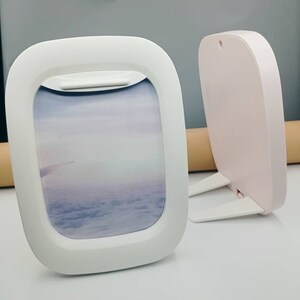 K&ouml;nnte beinhalten: Zwei Flugzeugfenster-Bilderrahmen in Wei&szlig; und Rosa mit St&auml;nder. Die Rahmen sind aus Kunststoff und haben ein realistisches Flugzeugfenster-Design. Der wei&szlig;e Rahmen hat ein Bild von Wolken und Himmel.