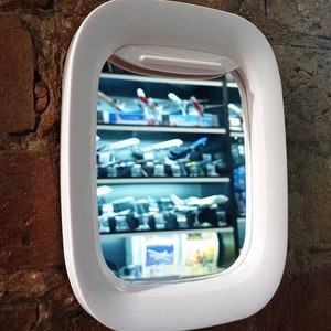 May include: White plastic airplane window mirror. The mirror is shaped like an airplane window and has a white frame. The mirror is reflecting a shelf of model airplanes.