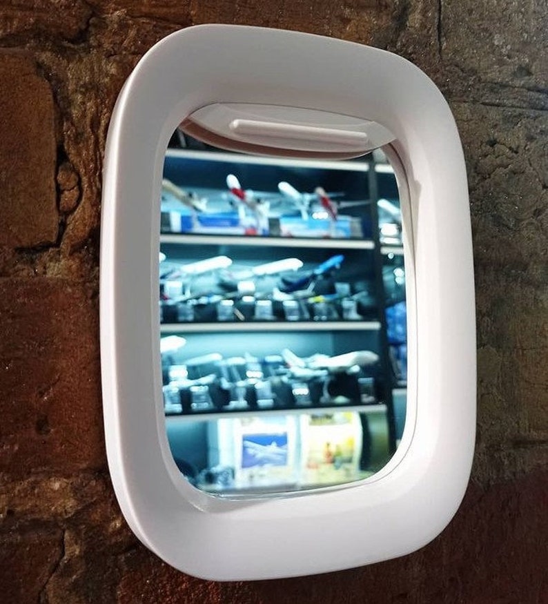 May include: White plastic airplane window mirror. The mirror is shaped like an airplane window and has a white frame. The mirror is reflecting a shelf of model airplanes.