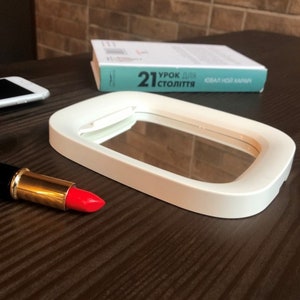 May include: White oval-shaped makeup mirror with a white frame and a clear glass surface. The mirror is sitting on a wooden table with a red lipstick and a cell phone.