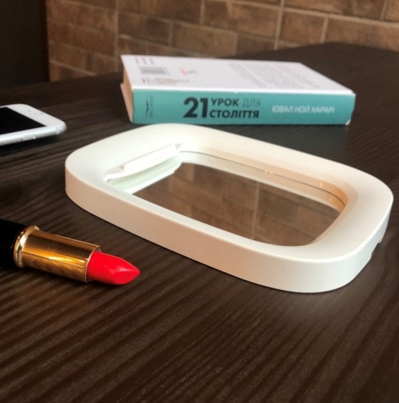 May include: White oval-shaped makeup mirror with a white frame and a clear glass surface. The mirror is sitting on a wooden table with a red lipstick and a cell phone.