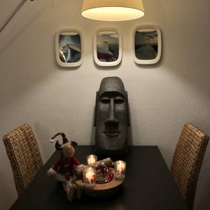 K&ouml;nnte beinhalten: Eine dunkelgraue Steinskulptur eines Osterinselkopfes steht auf einem schwarzen Tisch mit vier St&uuml;hlen. An der Wand hinter der Skulptur befinden sich drei wei&szlig;e gerahmte Bilder. Die Bilder zeigen Flugzeuge. Auf dem Tisch vor der Skulptur stehen vier brennende Kerzen.