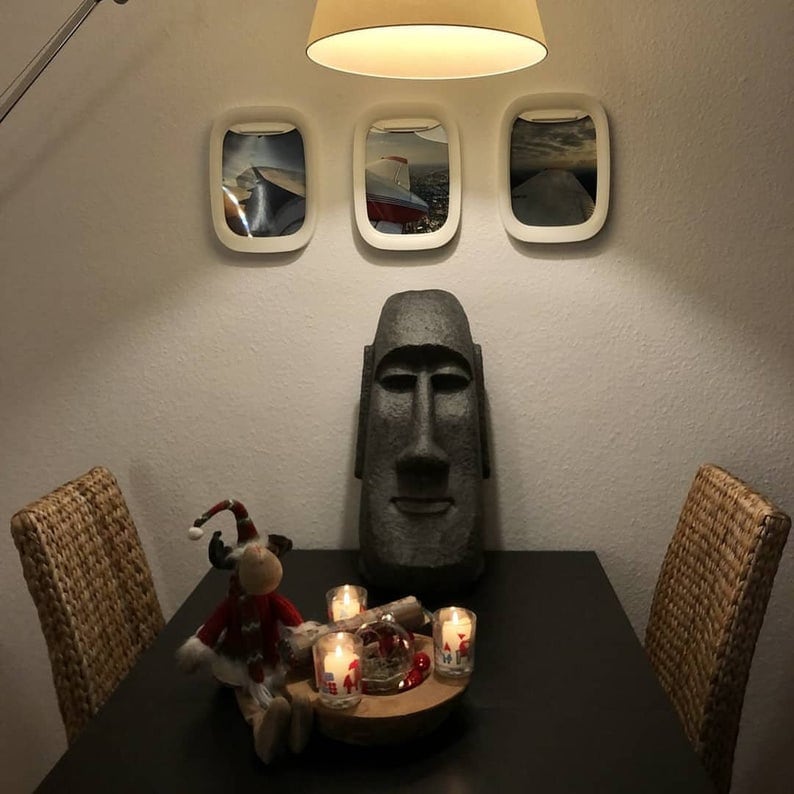 K&ouml;nnte beinhalten: Eine dunkelgraue Steinskulptur eines Osterinselkopfes steht auf einem schwarzen Tisch mit vier St&uuml;hlen. An der Wand hinter der Skulptur befinden sich drei wei&szlig;e gerahmte Bilder. Die Bilder zeigen Flugzeuge. Auf dem Tisch vor der Skulptur stehen vier brennende Kerzen.