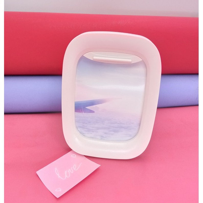 K&ouml;nnte beinhalten: Wei&szlig;er Flugzeugfenster-Bilderrahmen mit einem Bild von Wolken und einem Fl&uuml;gel. Der Rahmen steht auf einer rosa Oberfl&auml;che mit einer rosa Notiz, auf der "love" steht.