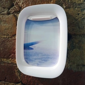 K&ouml;nnte beinhalten: Wei&szlig;er Flugzeugfensterrahmen aus Kunststoff mit einem Foto eines Fl&uuml;gels und Wolken. Das Foto ist eine Ansicht aus einem Flugzeugfenster.