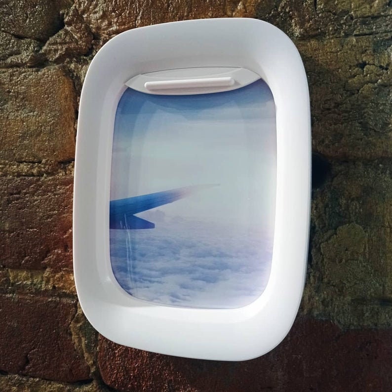 K&ouml;nnte beinhalten: Wei&szlig;er Flugzeugfensterrahmen aus Kunststoff mit einem Foto eines Fl&uuml;gels und Wolken. Das Foto ist eine Ansicht aus einem Flugzeugfenster.