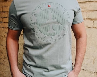 Può includere: Una t-shirt verde chiaro con un design a bussola che presenta una silhouette di aereo. La bussola ha numeri da 0 a 36 e le lettere N, E, S e W.